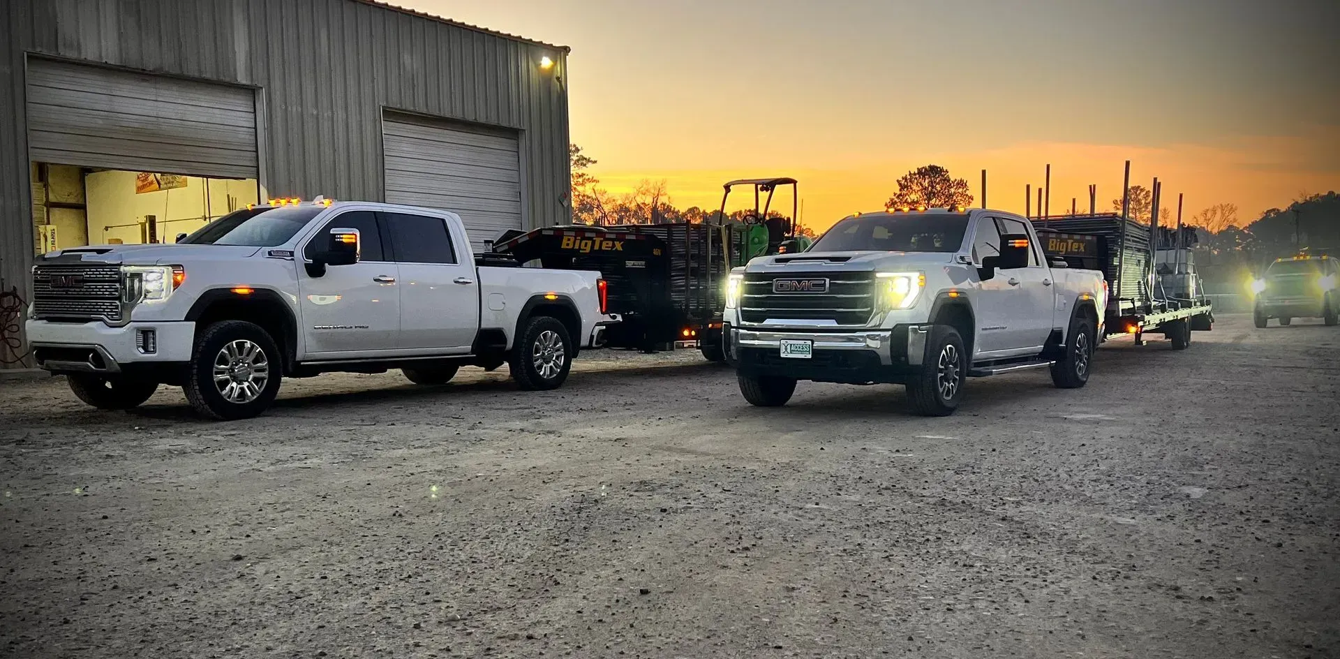 Two white pickup trucks parked outside a metal building at sunset with headlights on