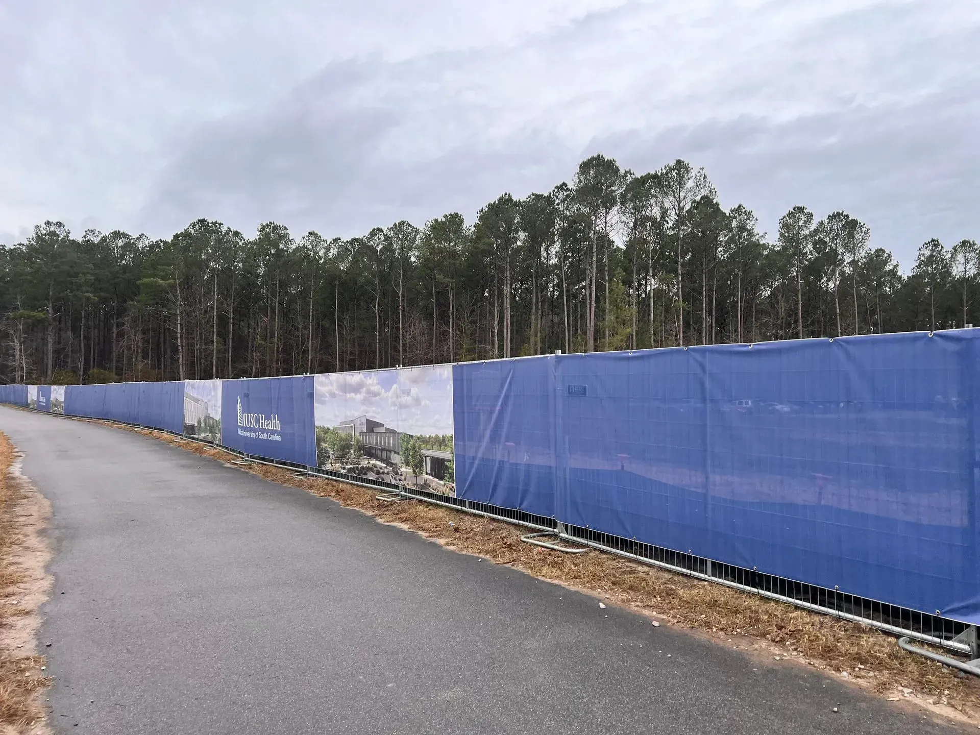 Blue construction fence along a paved road beside a wooded area under a cloudy sky