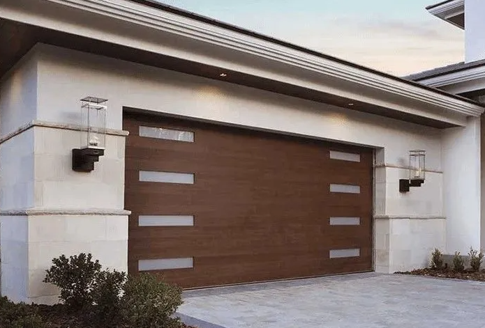 Brown garage door with rectangular glass panels, light-colored exterior wall, and wall-mounted lights.