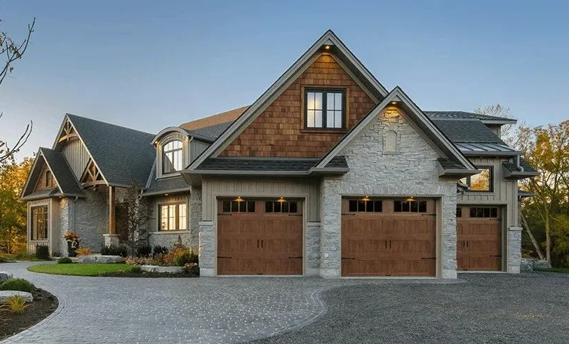 Two-story house with stone and brown exterior, three garage doors, and a paved driveway.
