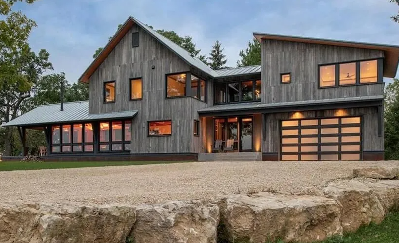 Modern gray wood house with large windows, gravel driveway, and stone border.