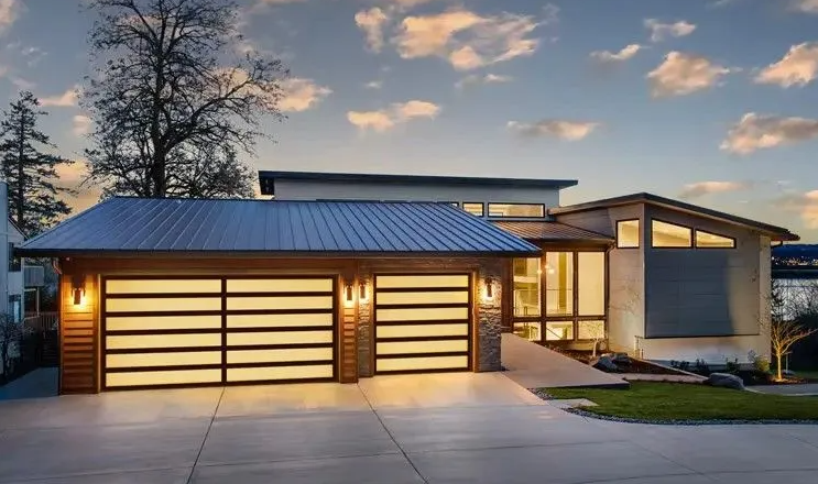 Modern home with two-car garage, lit garage doors, wood and stone facade, and a concrete driveway at dusk.