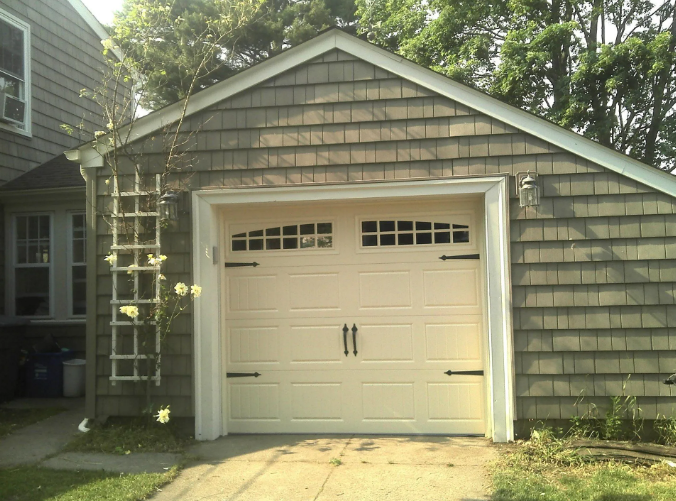 Beige garage door with black accents and trim, brown shingles, and a climbing plant.