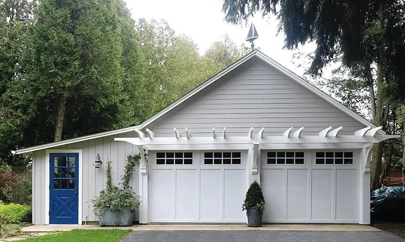 Two-car garage with white doors and a blue side door. Grey siding and a pergola above the garage doors.