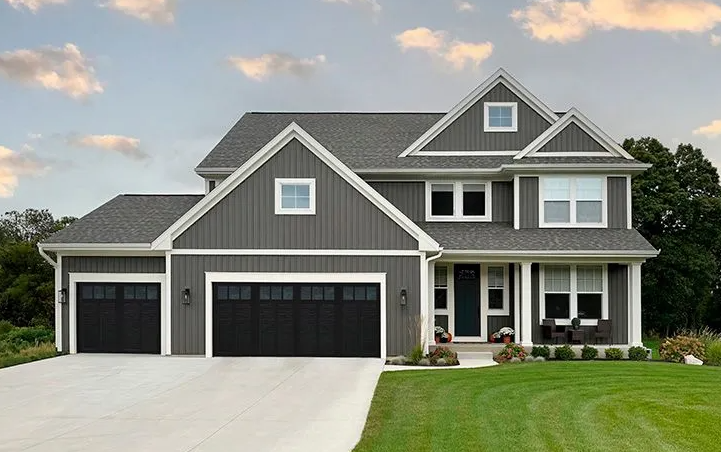 Gray two-story house with black garage doors and white trim, set on a green lawn with a cloudy sky.