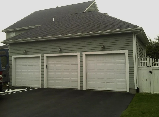 Three-car garage with white doors, gray siding, and a dark asphalt driveway. A section of white fence is on the right.