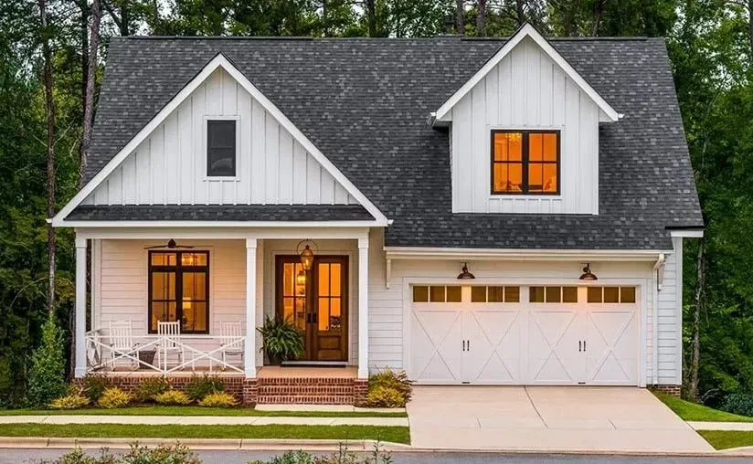 White farmhouse-style home with black roof, garage, and front porch, set against a backdrop of green trees.