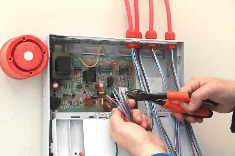 Person cutting wires with pliers inside an open electrical box. Red alarm bell on the left.