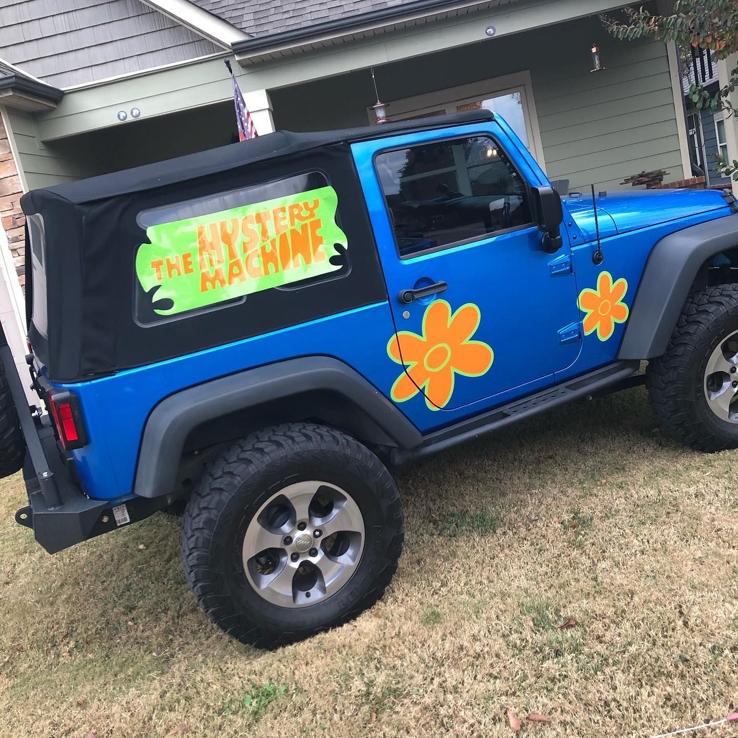 Blue Jeep Wrangler modified to resemble the Mystery Machine from Scooby-Doo, parked on grass with house in background.