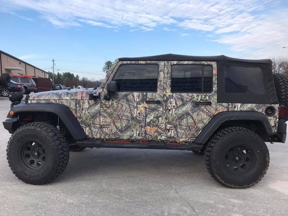 Camouflage Jeep Wrangler on a paved lot with black wheels, a black soft top, and tinted windows.