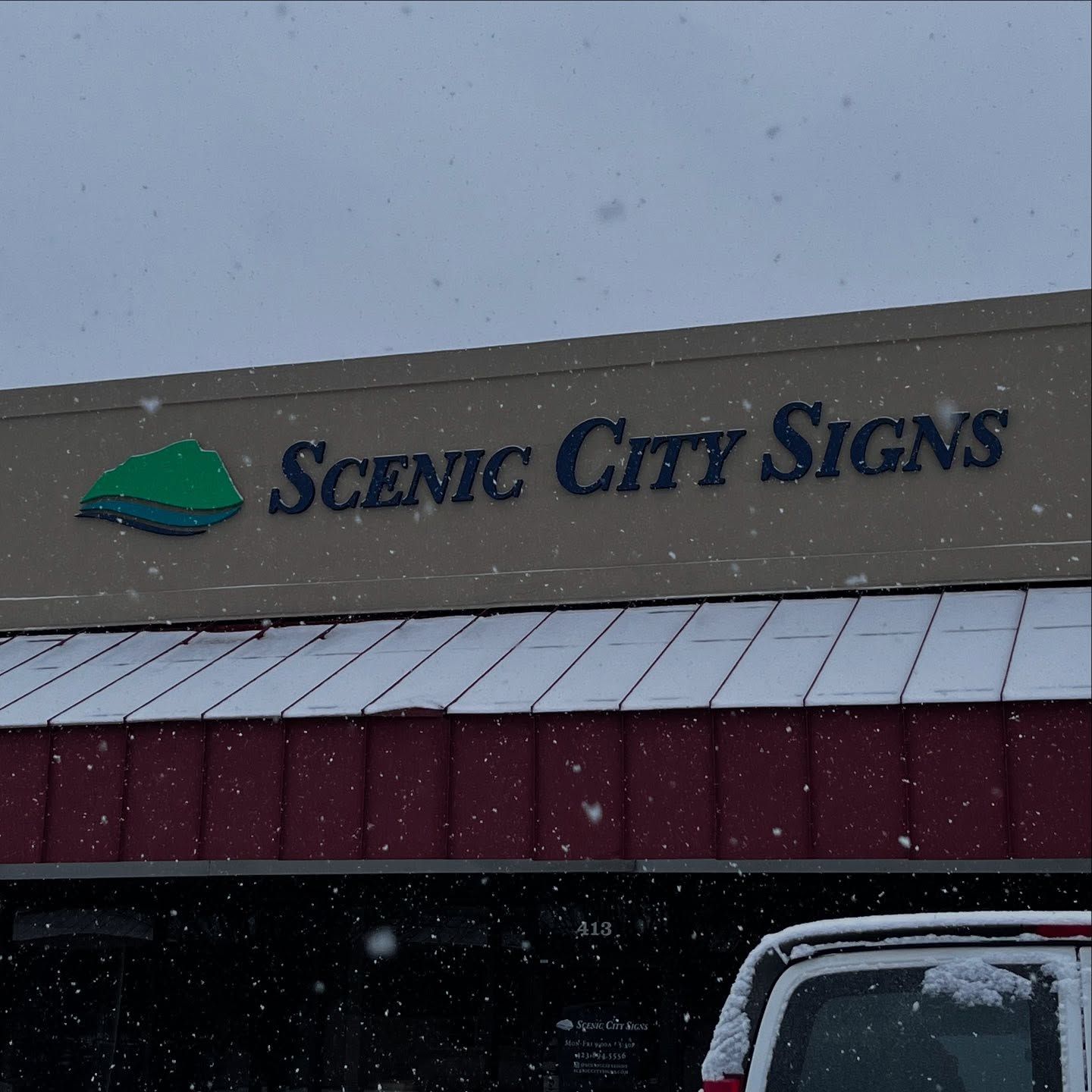 Scenic City Signs storefront with snow falling, on a cloudy day.