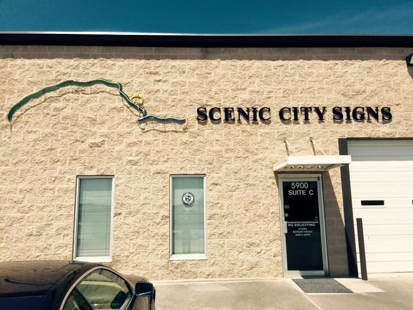 Scenic City Signs business exterior; tan brick, brown text, green and yellow river graphic, white garage door.