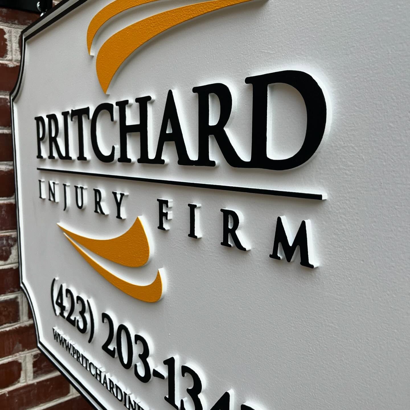 Sign for Pritchard Injury Firm, white with black and orange text and logo, on a brick wall.