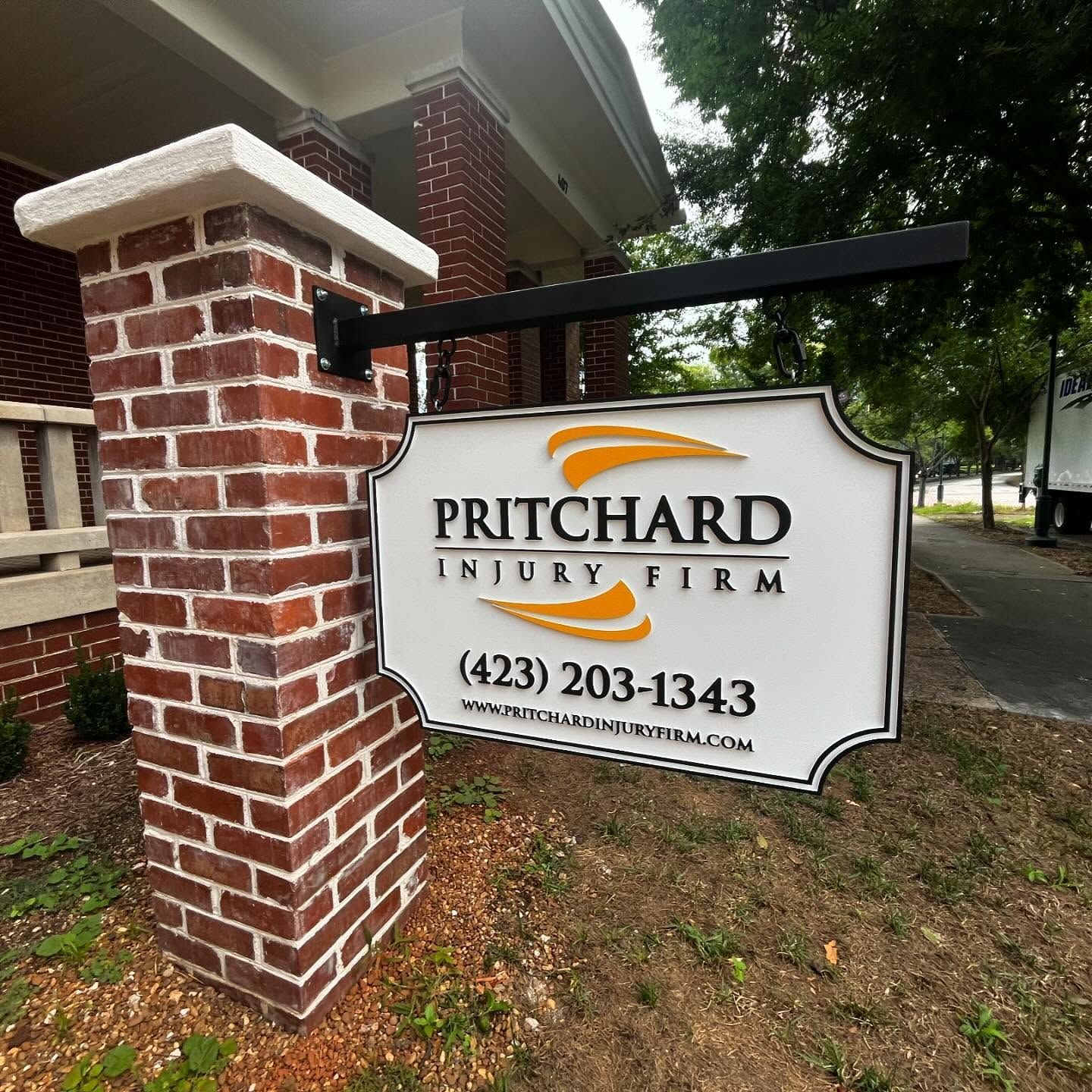 Sign for Pritchard Injury Firm on a brick pillar, featuring the firm's name, logo, phone number, and website.