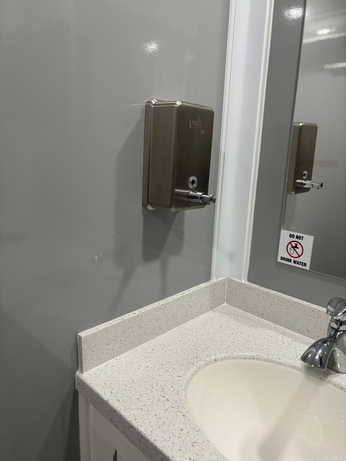 Soap dispenser on a gray wall above a countertop with a sink, in a restroom.