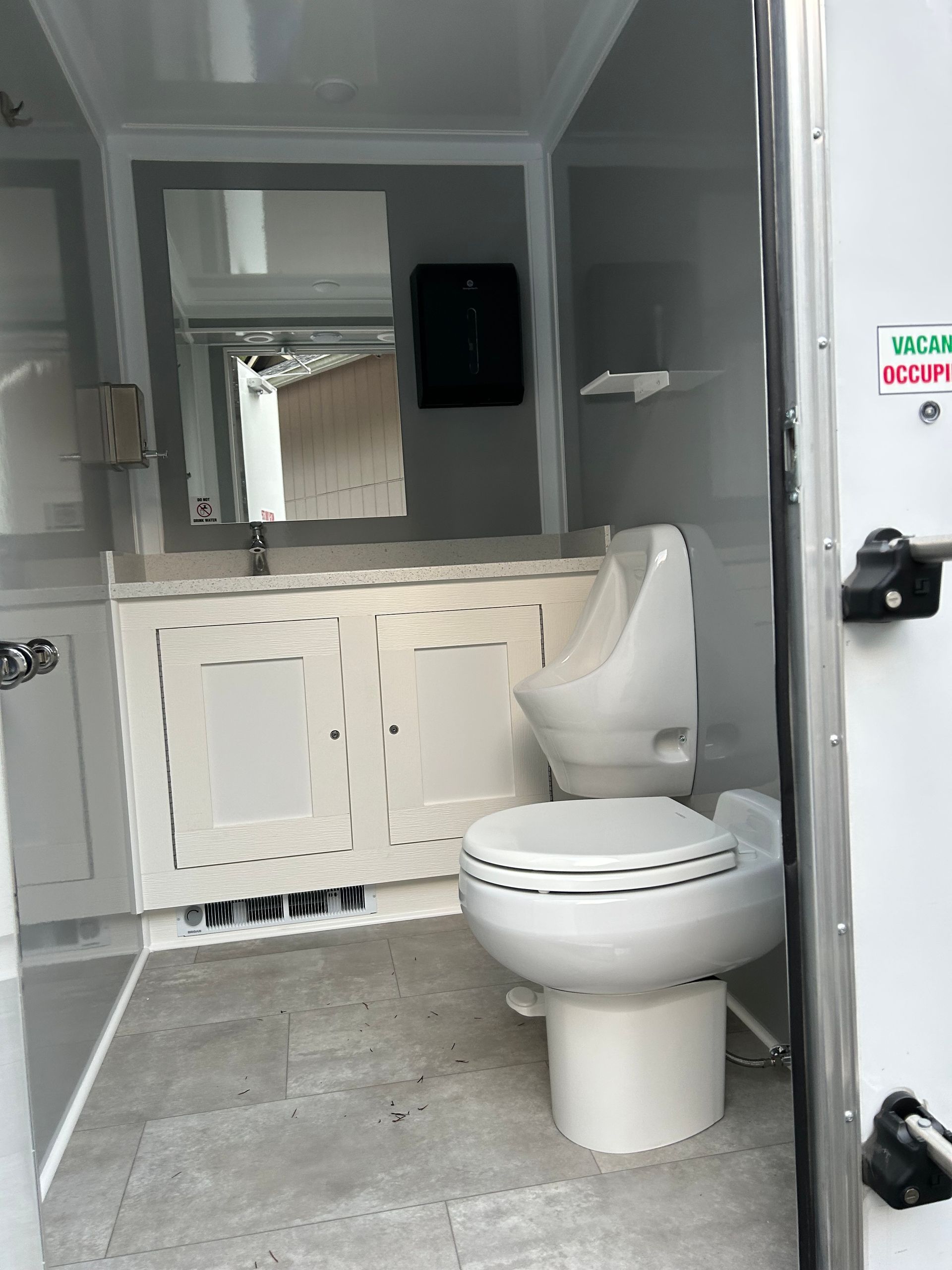 Interior of a portable restroom: white toilet, sink, and mirror. Gray floor and walls.