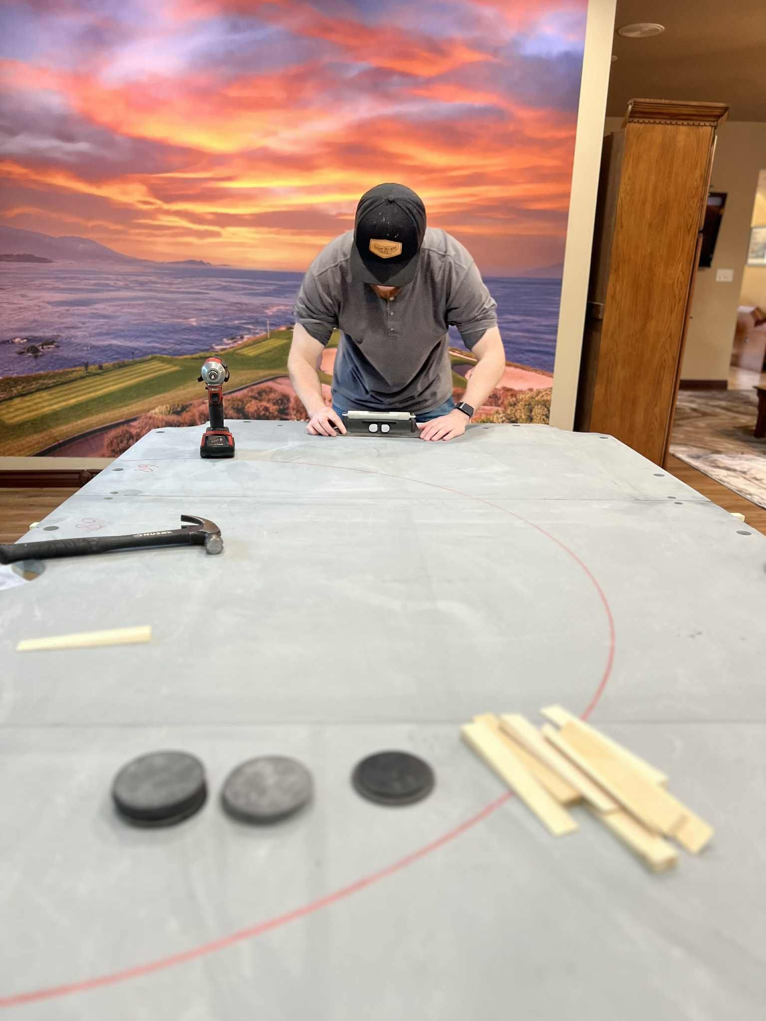 Person leaning over a worktable with tools and materials, in front of a sunset mural.