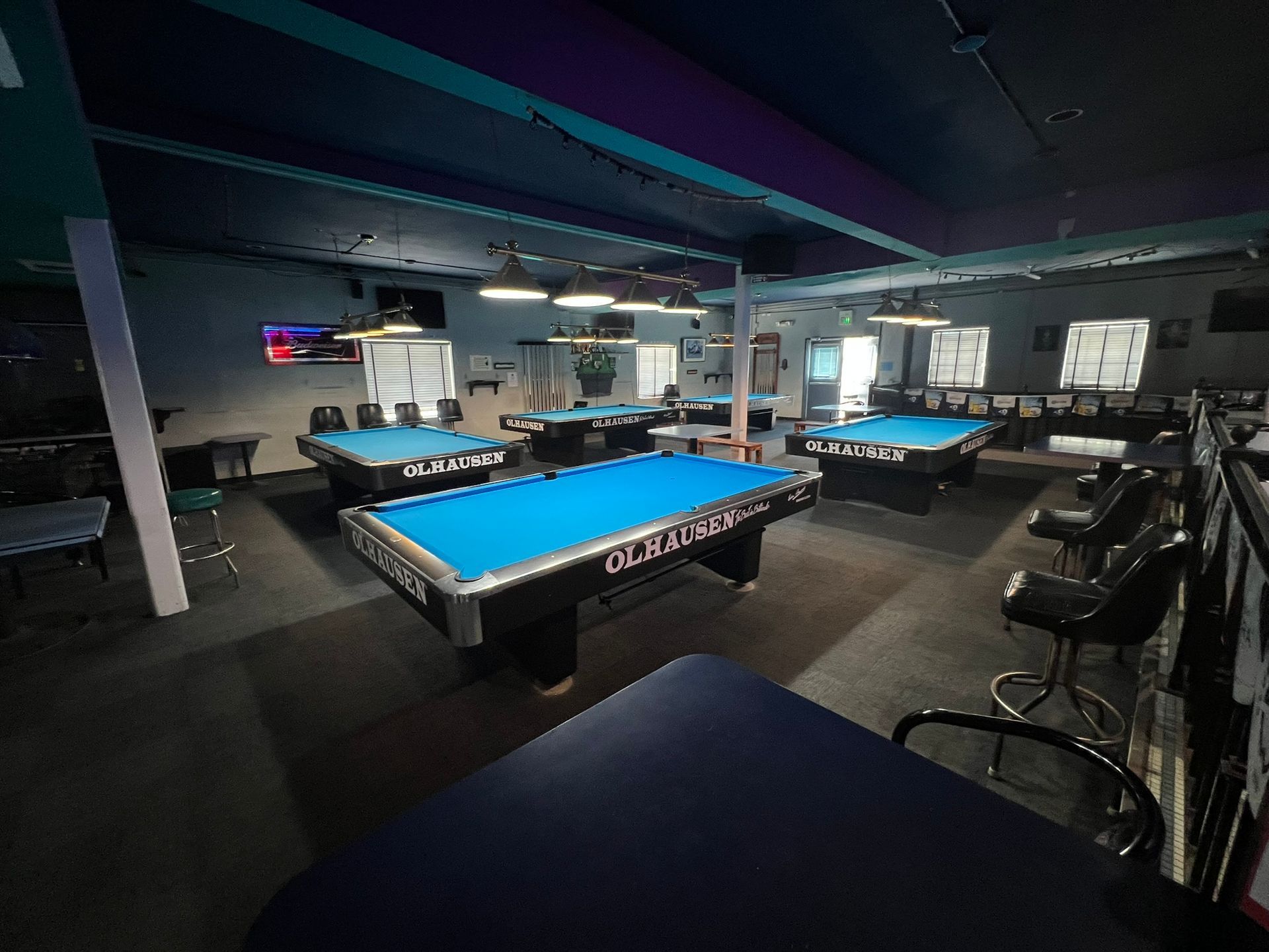 Dim billiards hall with several pool tables under low purple lighting.