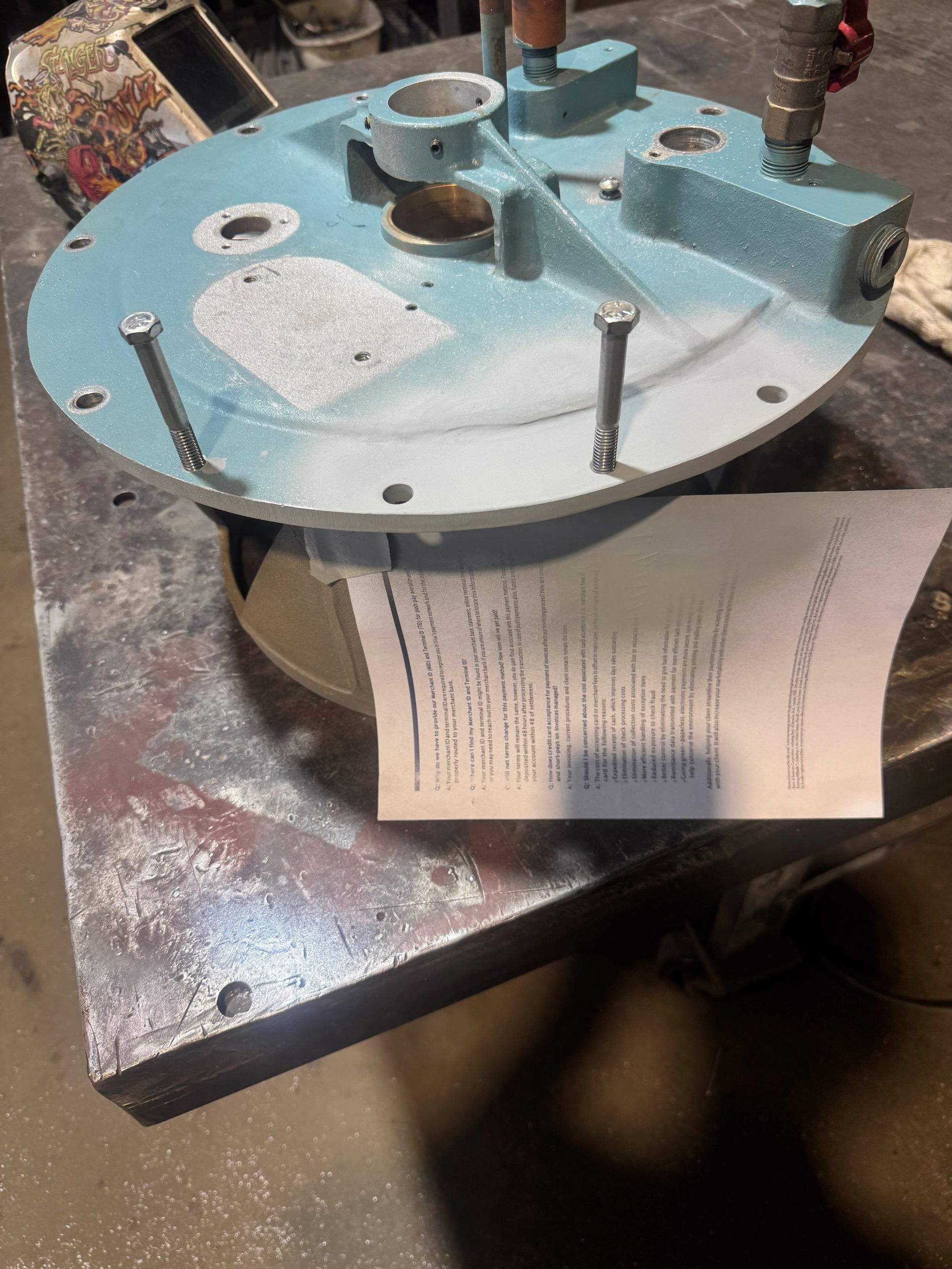 Blue metal mechanical assembly on a workbench, with bolts, pipes, and a paper tag attached.
