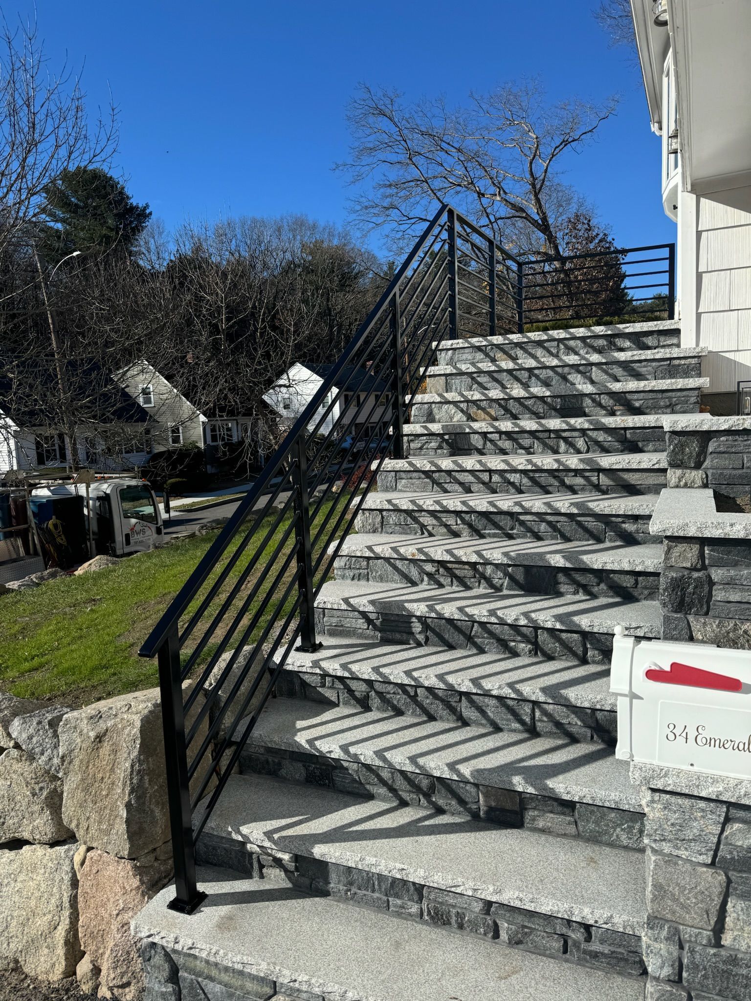 Outdoor stone staircase with black railing beside a house, viewed from below on a sunny day