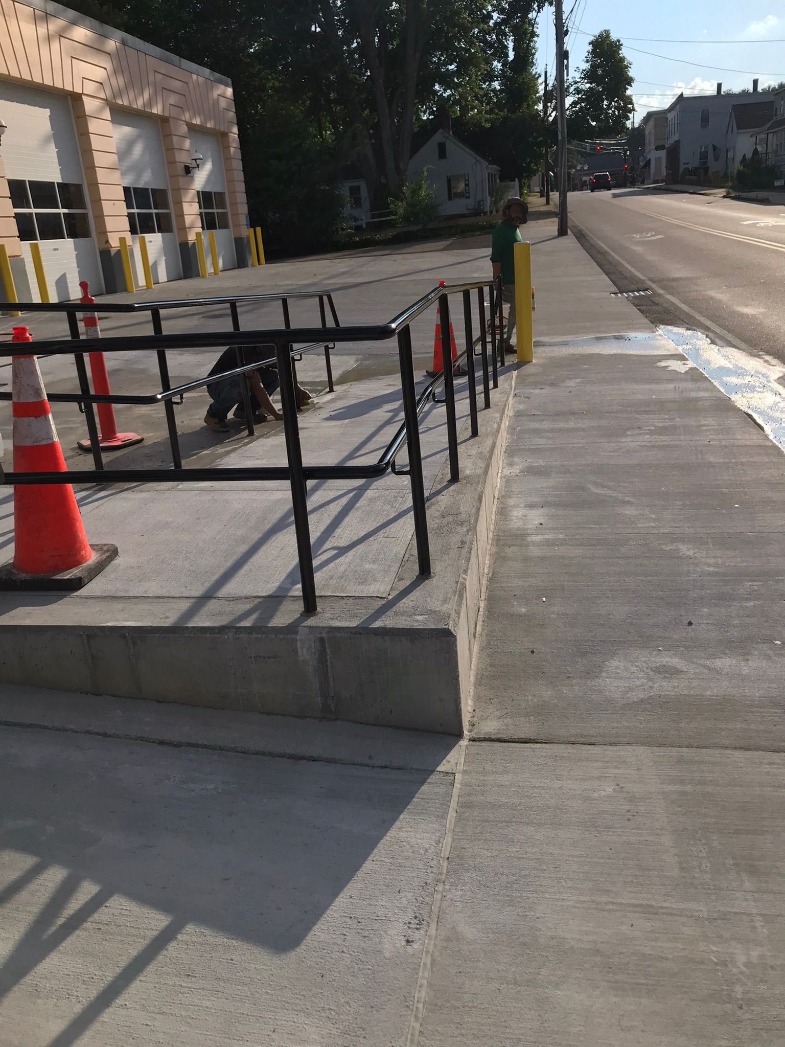 Street sidewalk ramp beside a building, with black railings and an orange traffic cone under construction.