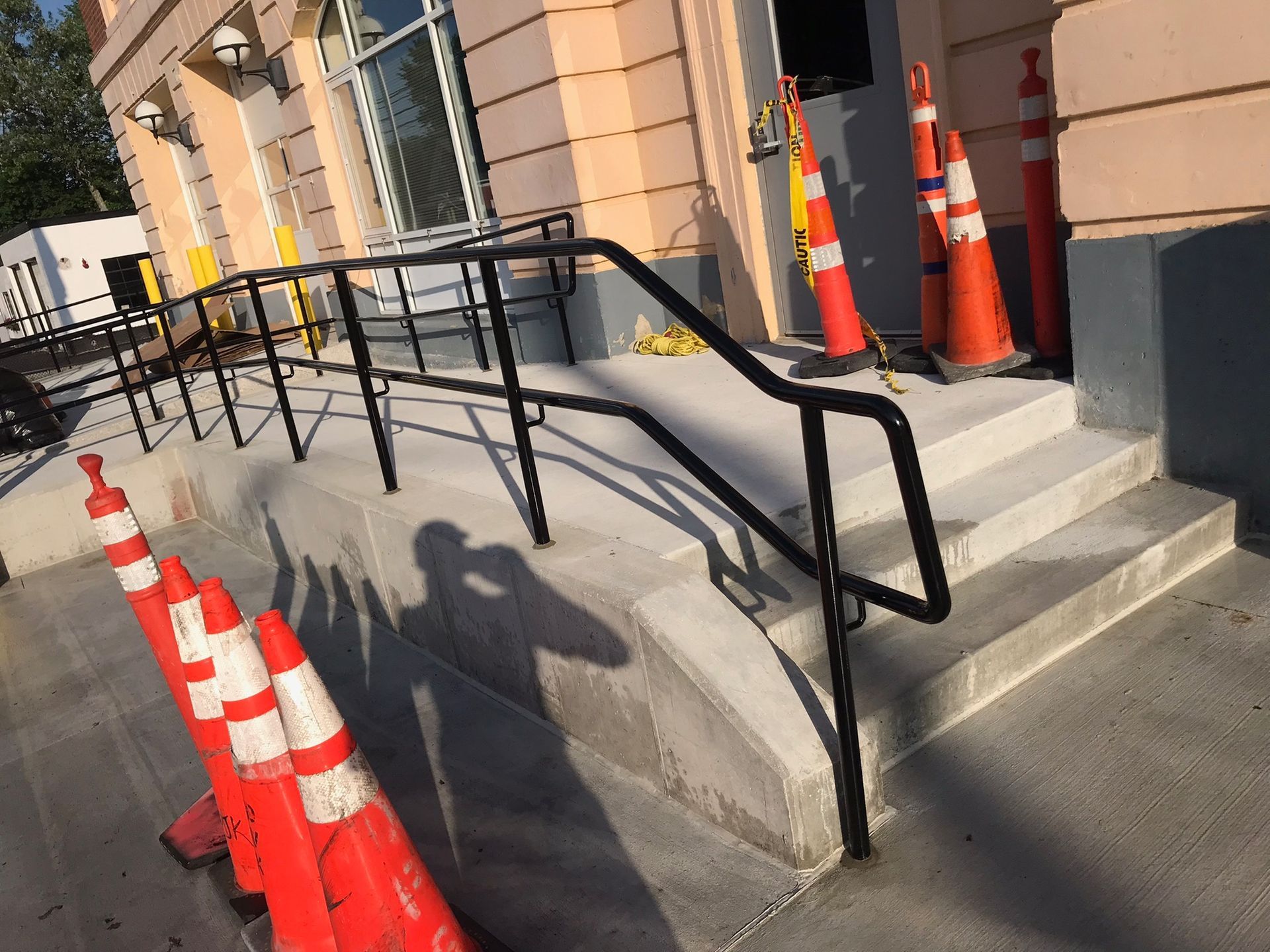 Building entrance ramp with black handrails, steps, and orange traffic cones on the sidewalk.
