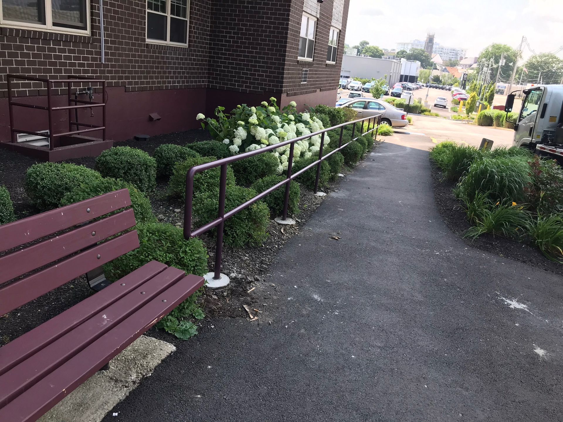 Walkway beside a brick building with benches, handrails, and shrubs, leading downhill to a neighborhood view