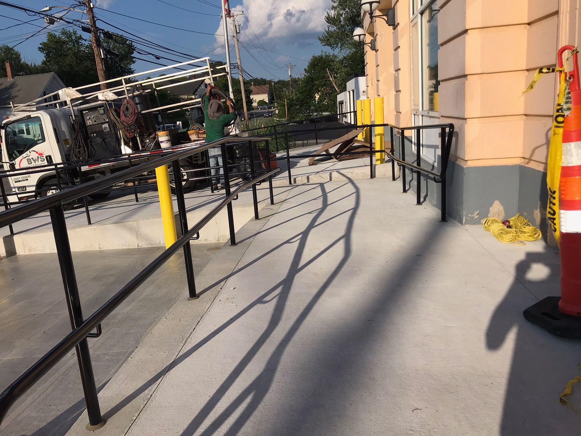 Ramped sidewalk beside a building with black railings and construction equipment nearby on a sunny day
