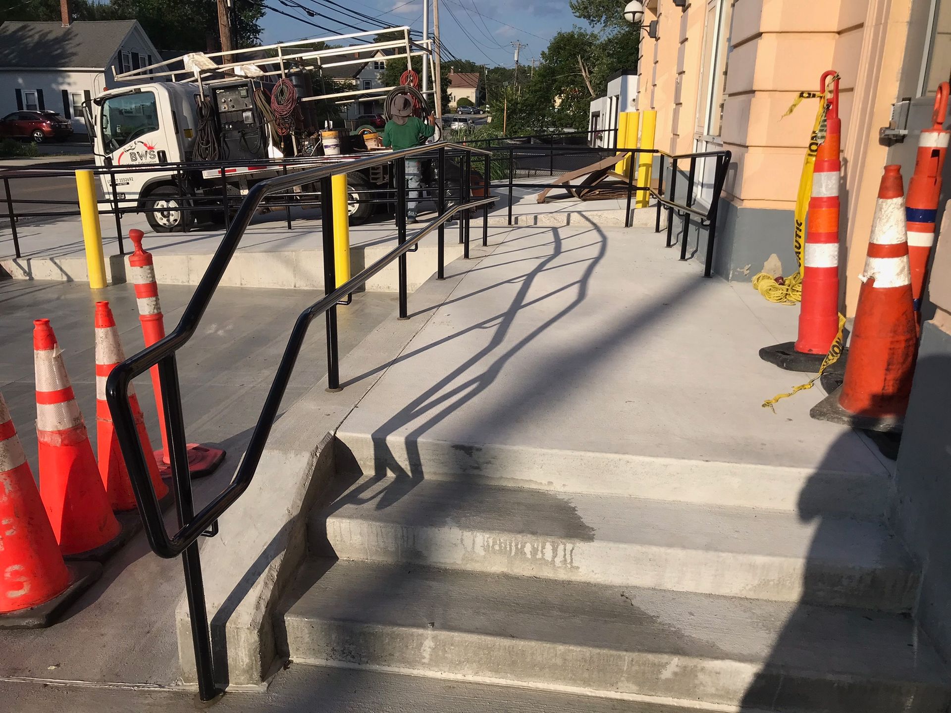 Accessible sidewalk ramp with black handrails beside a building, orange traffic cones, and steps in front