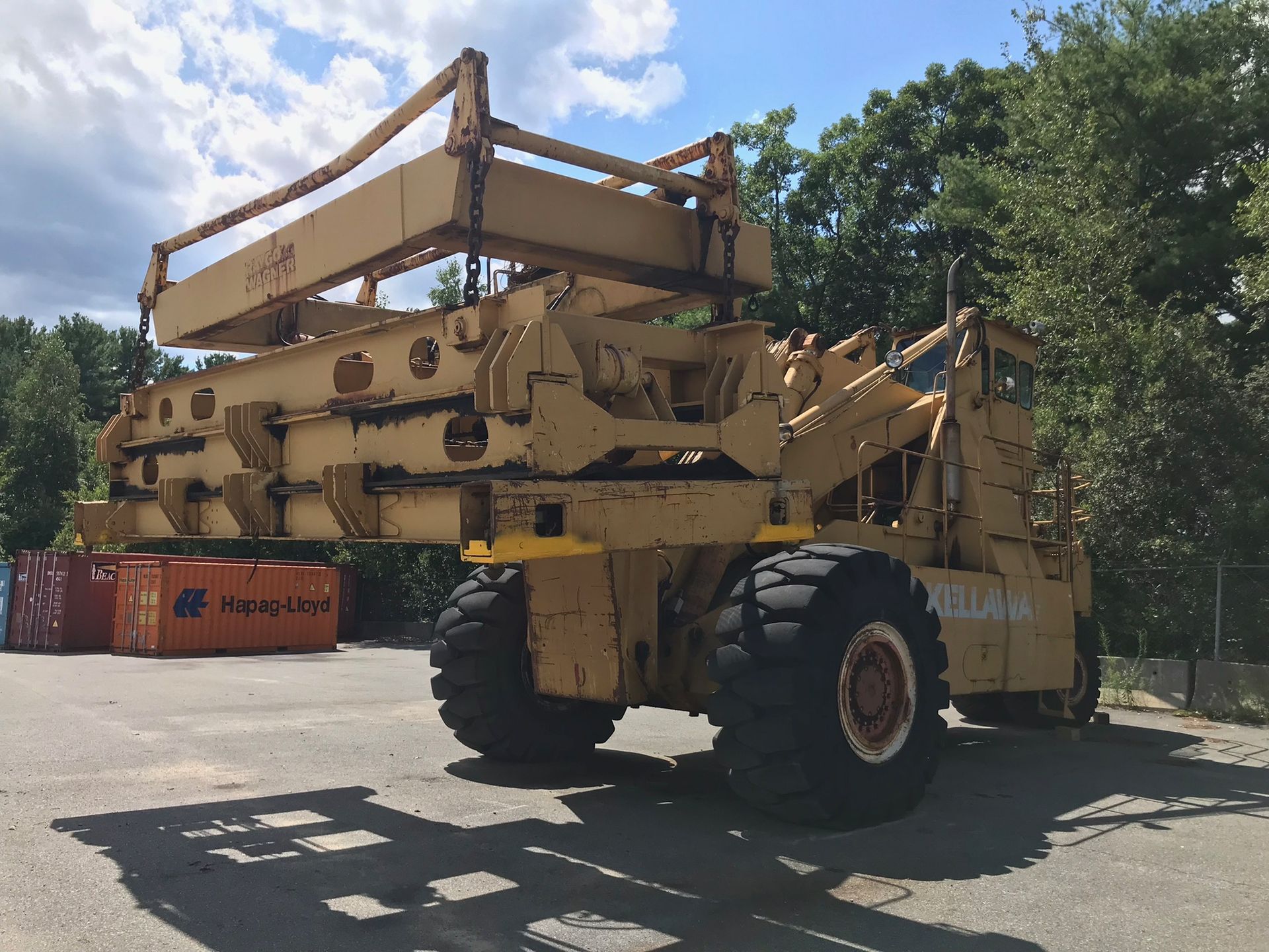 Large yellow industrial machine parked outdoors beside red shipping containers and trees.