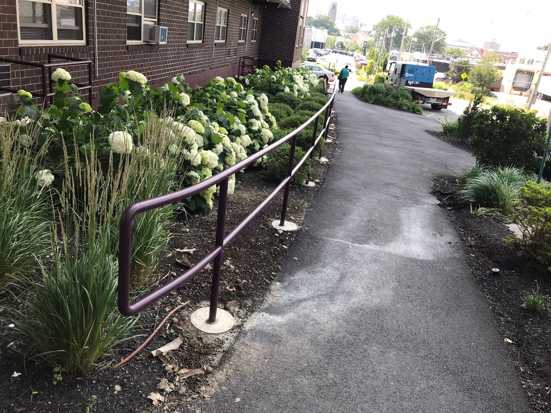 Curved sidewalk beside brick building with flower beds and a purple handrail, leading to a street
