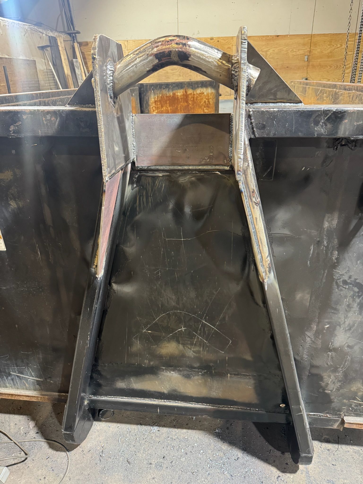 Black metal dumpster with open front and slanted supports in an indoor industrial setting