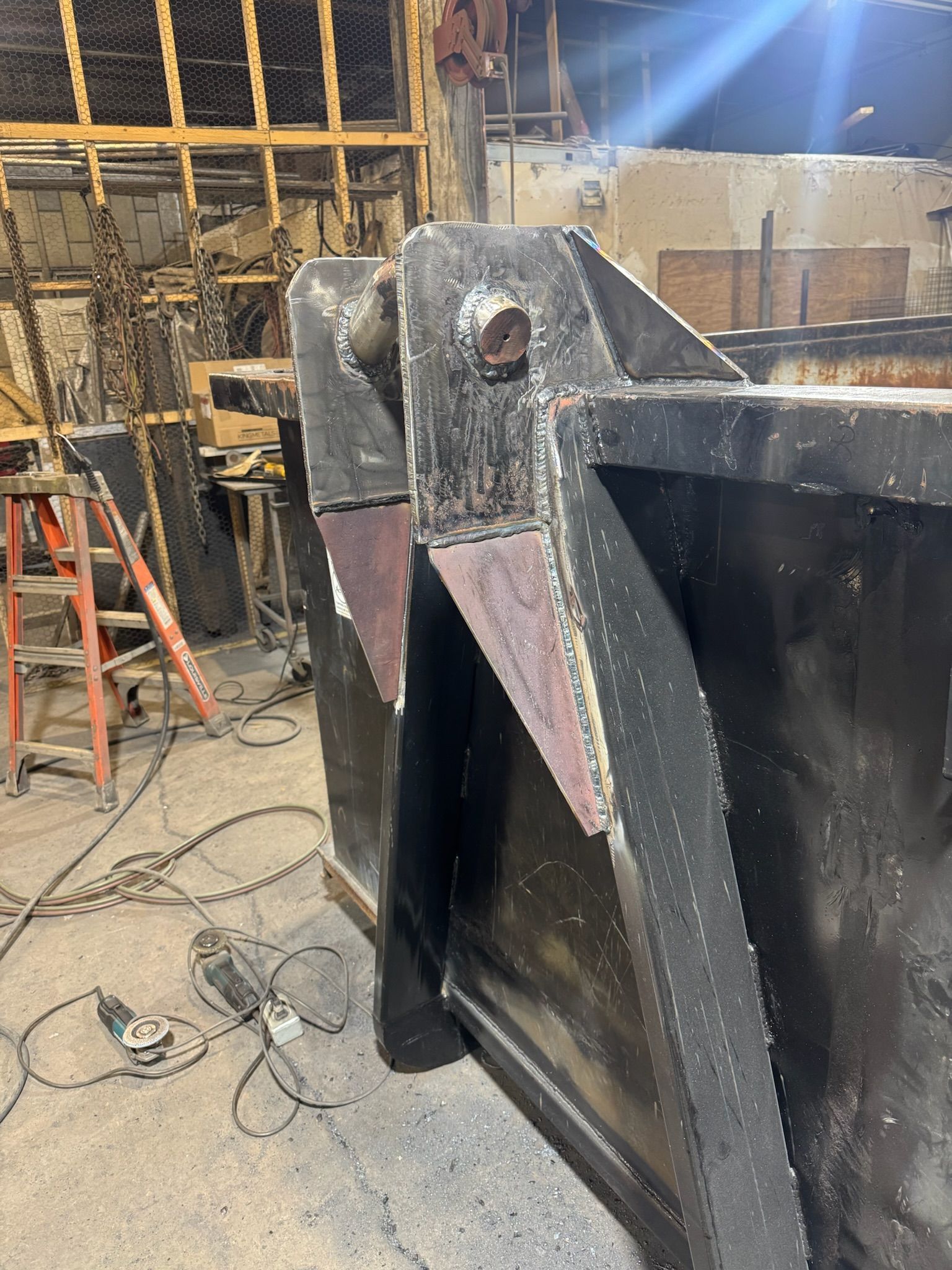 Large black steel beam in a workshop, with welds and a red ladder in the background.