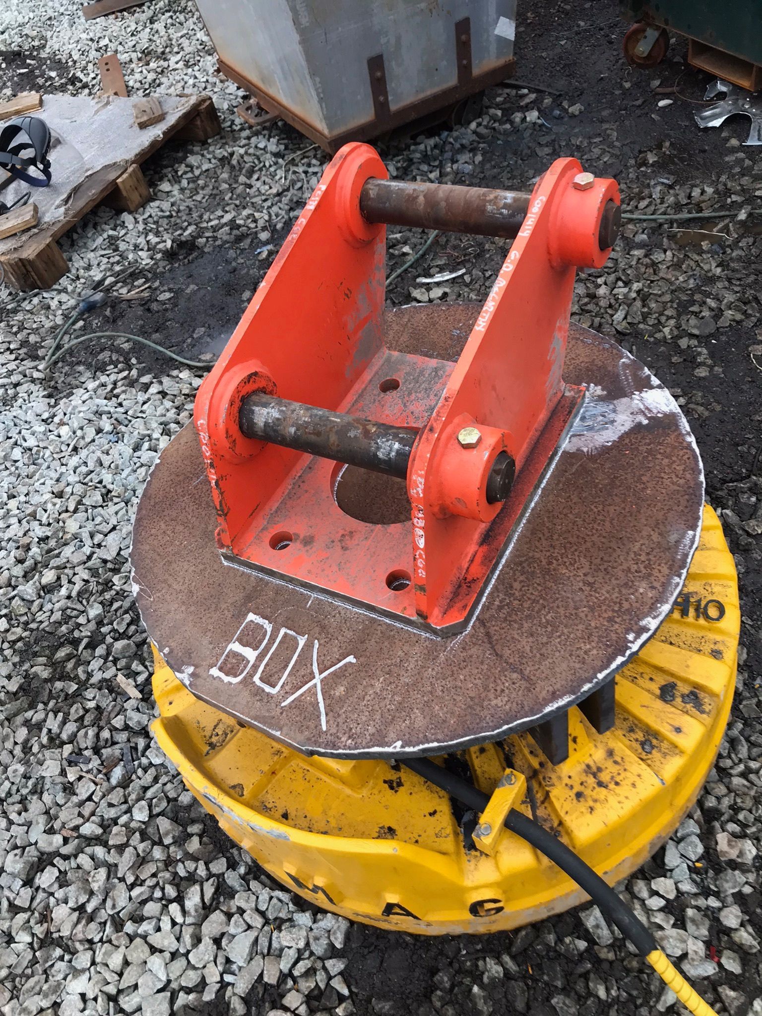 Red excavator quick coupler on a yellow construction attachment, resting on a metal plate outdoors.