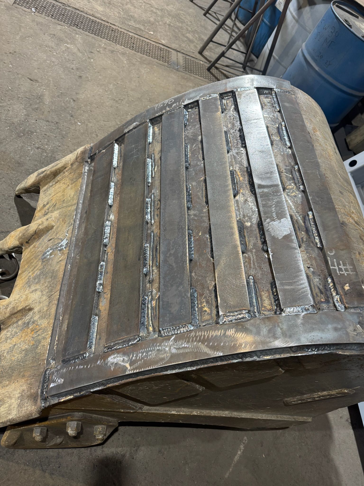 Rusty metal drum or barrel lid with slotted slats, viewed on a concrete floor near blue barrels.