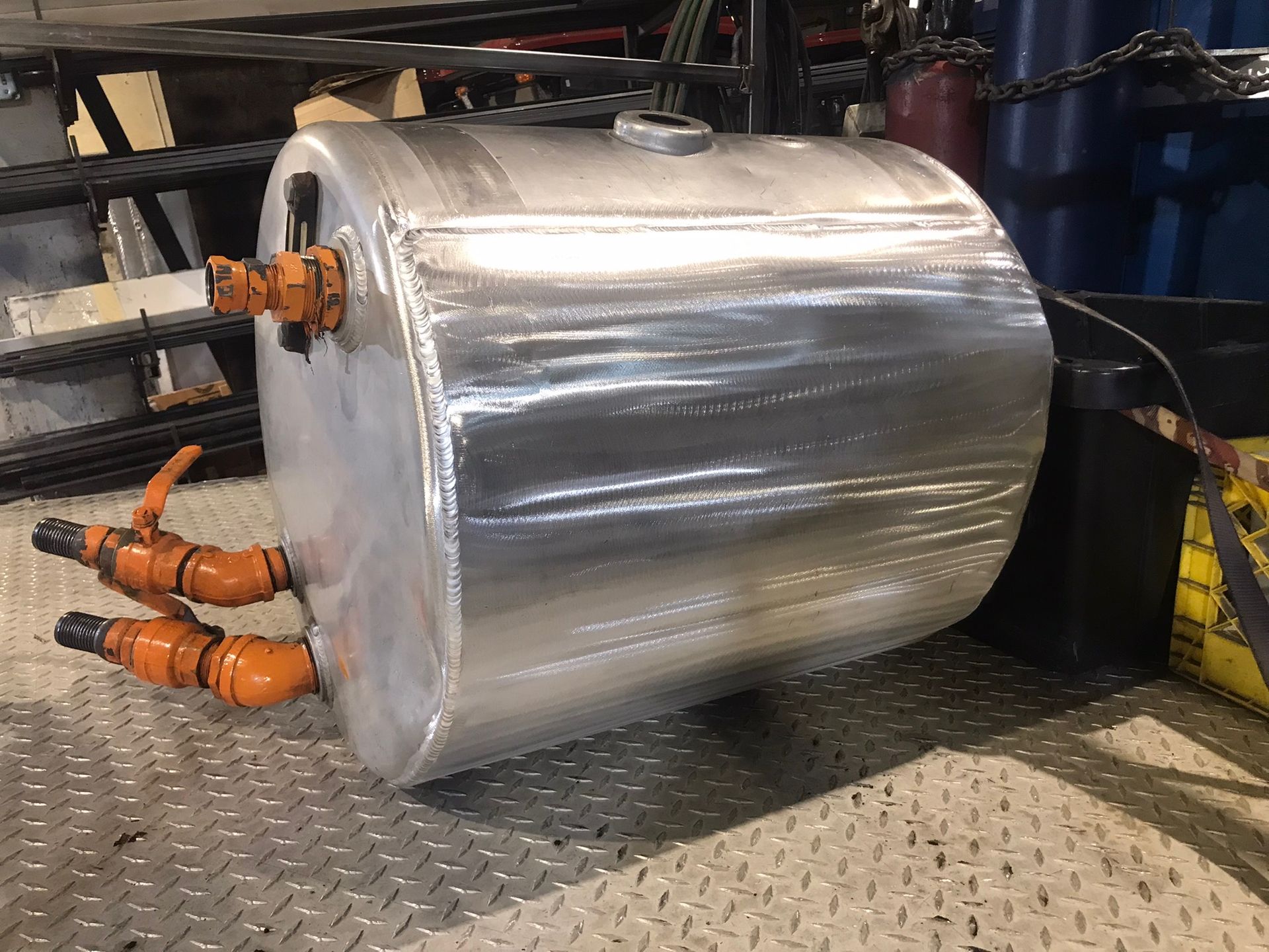 Large silver cylindrical tank with orange valves, resting on a metal floor in an industrial workshop