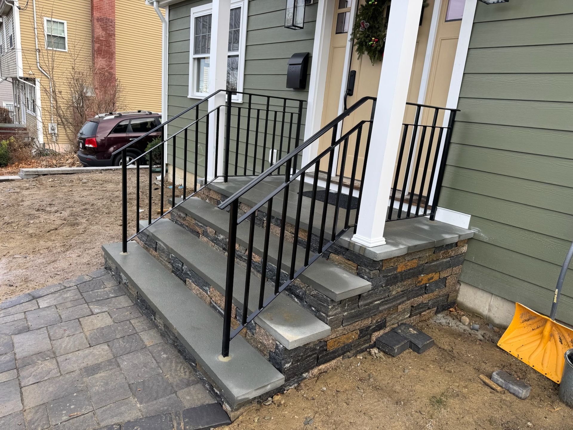Front steps and porch with black railings, gray stone steps, and a tan house exterior beside a paved walkway