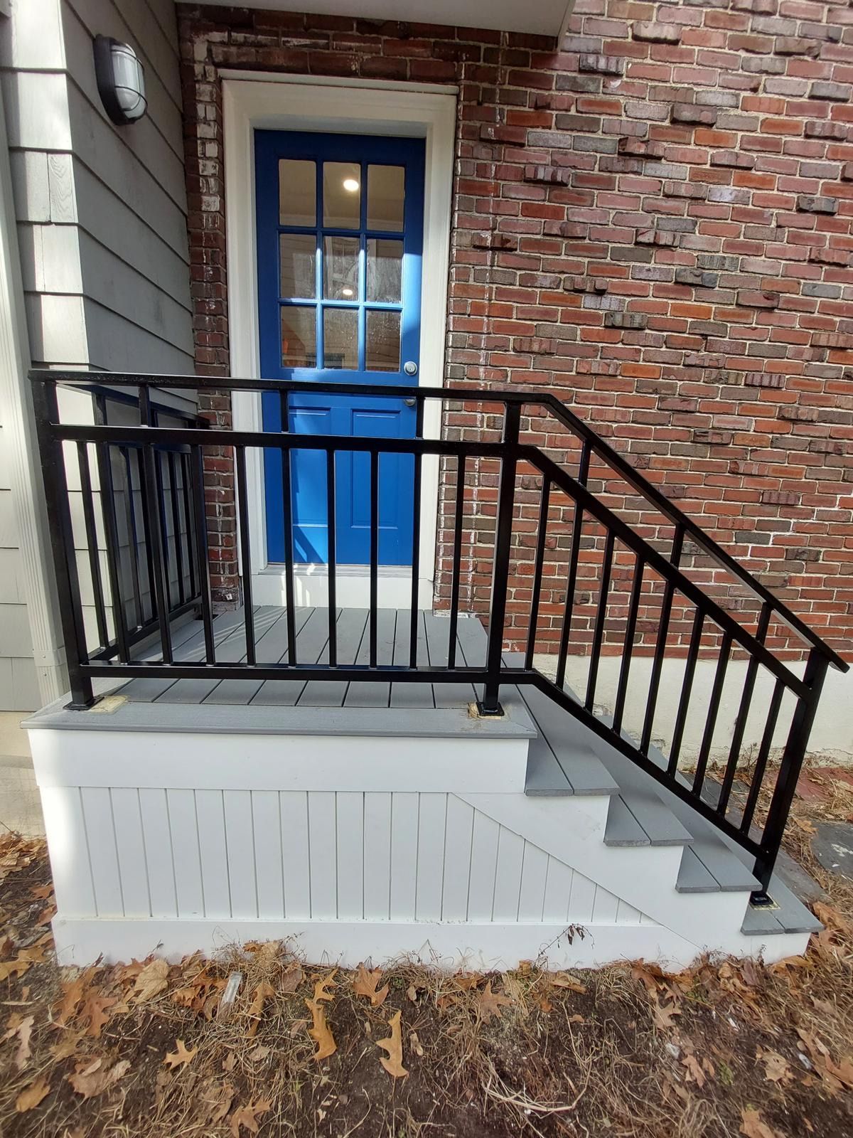 Front porch with blue door, brick wall, black metal railing, and white foundation steps covered in leaves