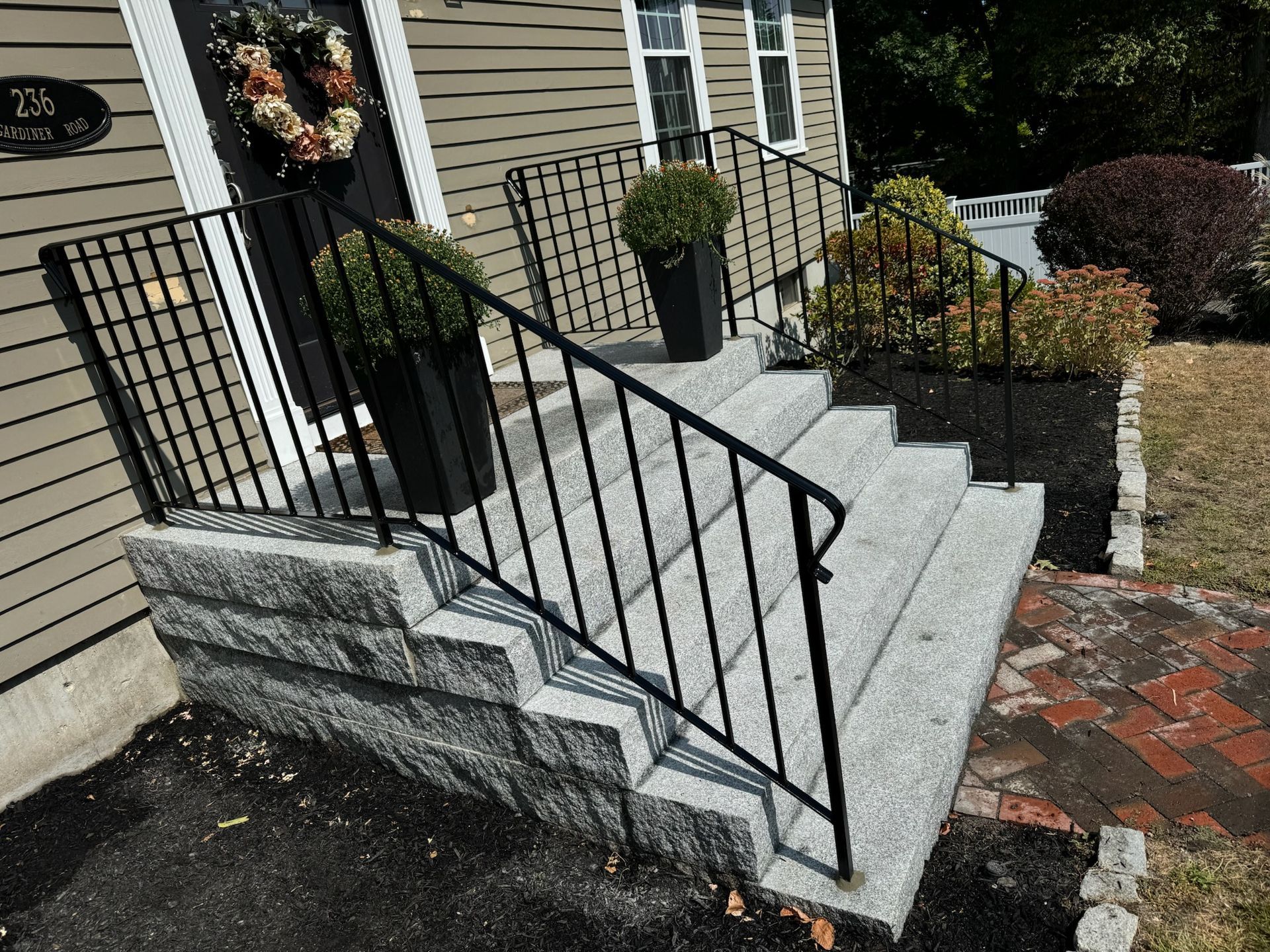 Front steps with black railing leading to a house entrance, beside a brick walkway and potted plants