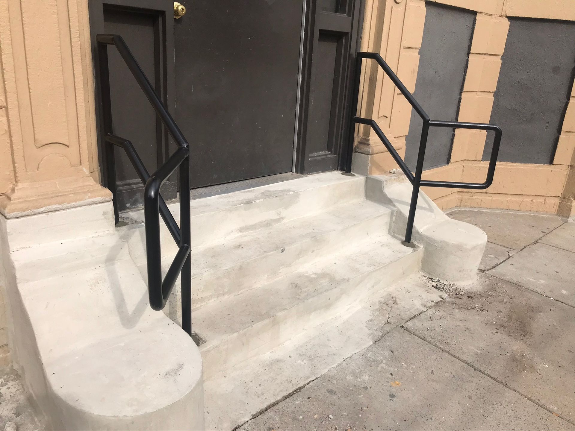 Concrete steps with black metal handrails leading to a dark doorway on a tan building.