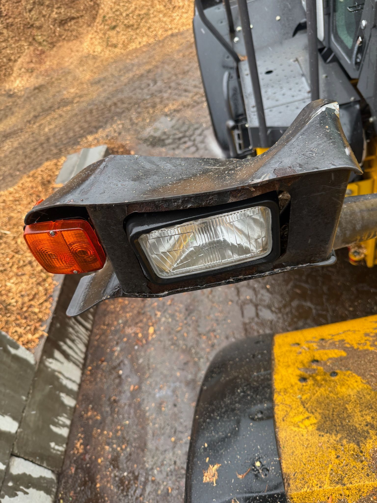 Close-up of a black vehicle’s front light assembly with a cracked clear lens and amber side marker, wet with dirt.