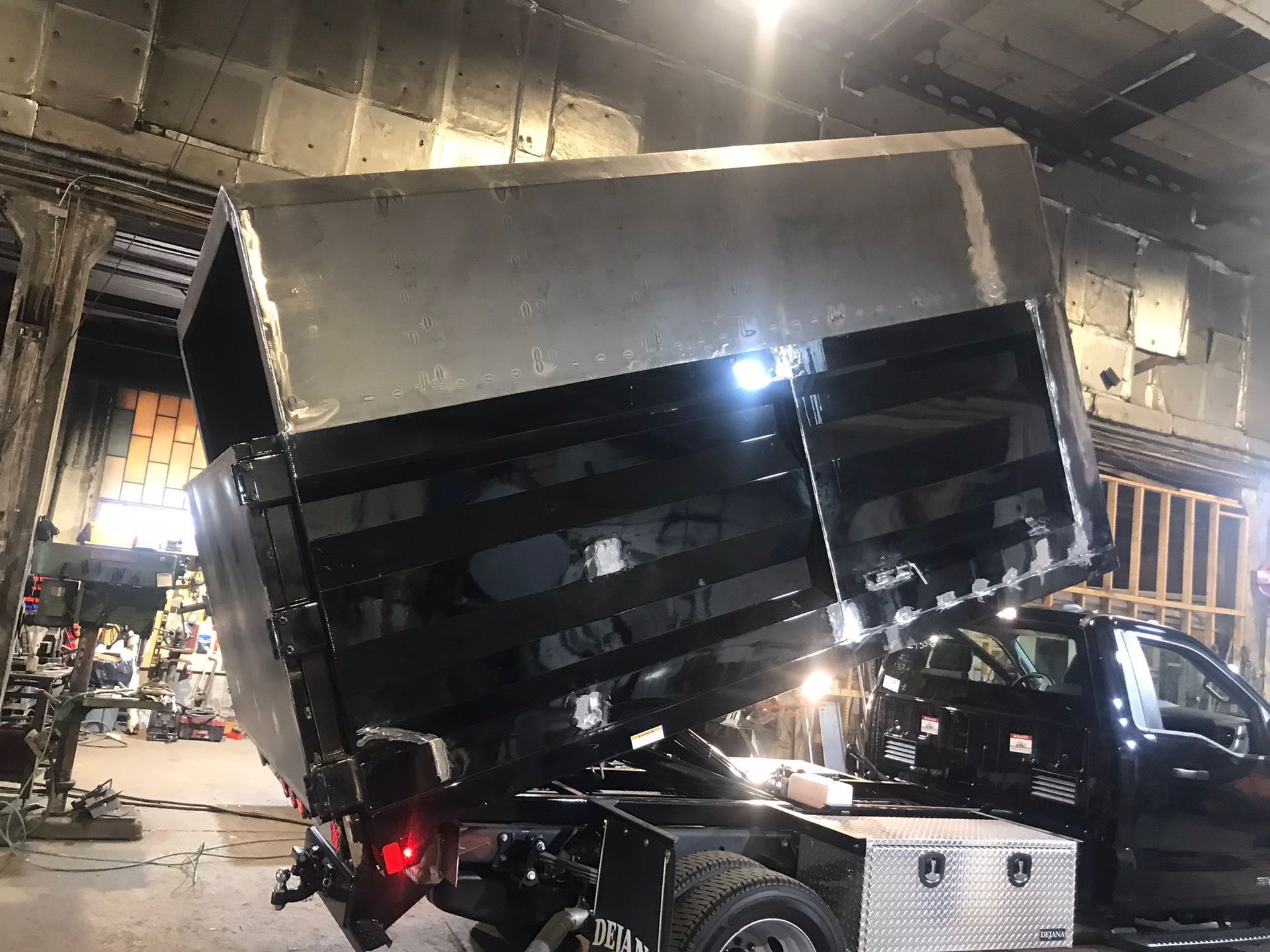 Large dump truck with raised bed inside a garage, rear lights on, tilted heavily to one side