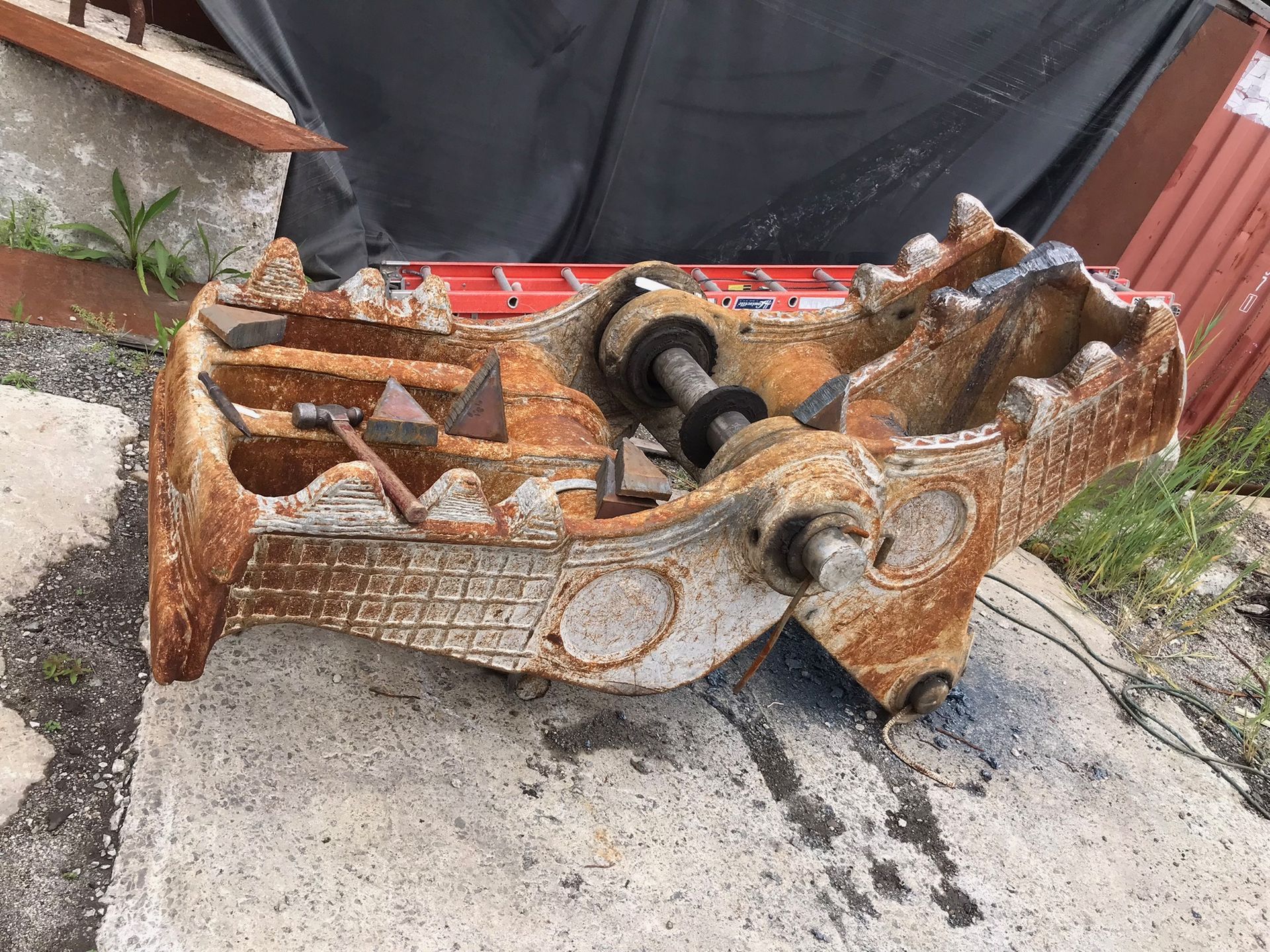 Rusty excavator bucket attachment lying on concrete beside a red wall and plants