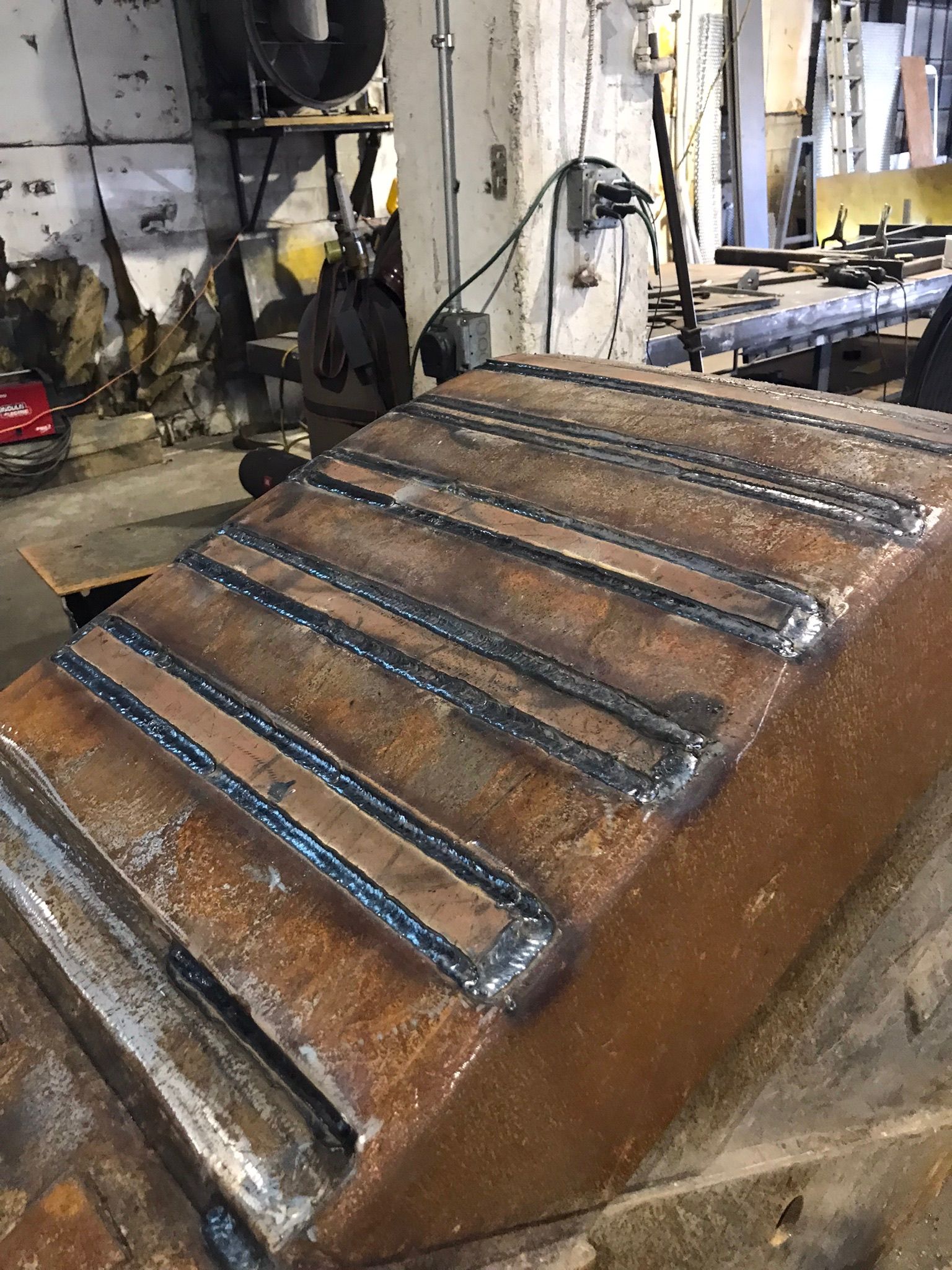 Rusty brown industrial metal chute with raised black bars in a workshop or factory setting