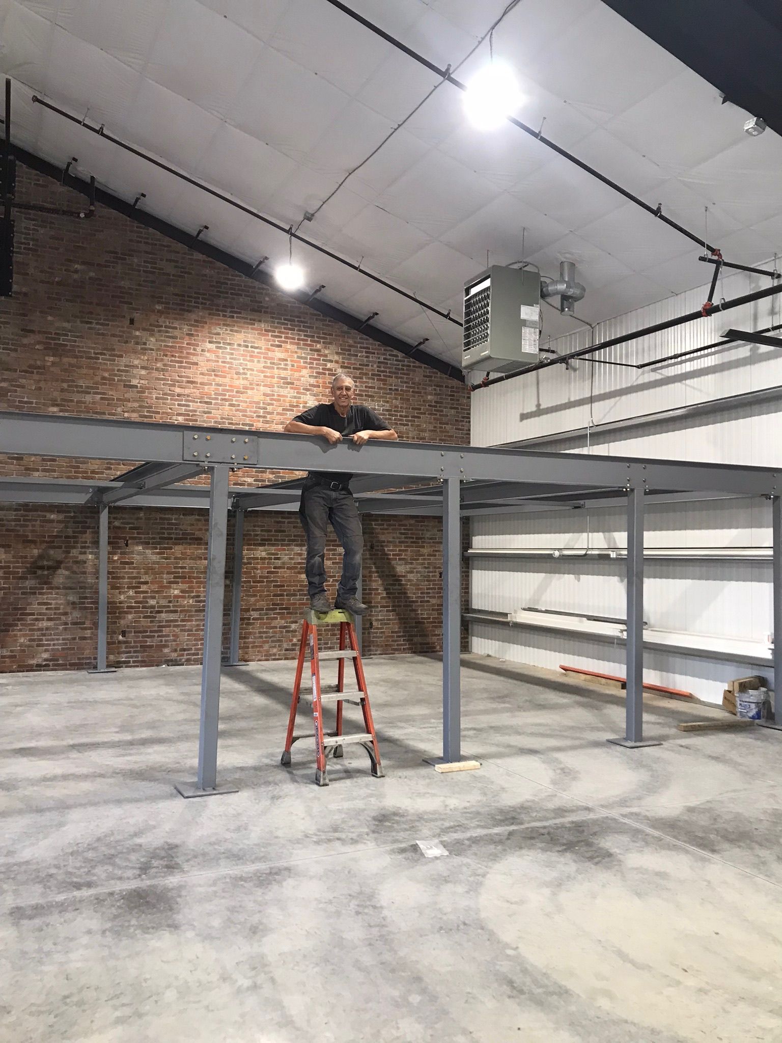 Worker on ladder assembling a metal mezzanine frame inside a warehouse with brick walls and high ceilings