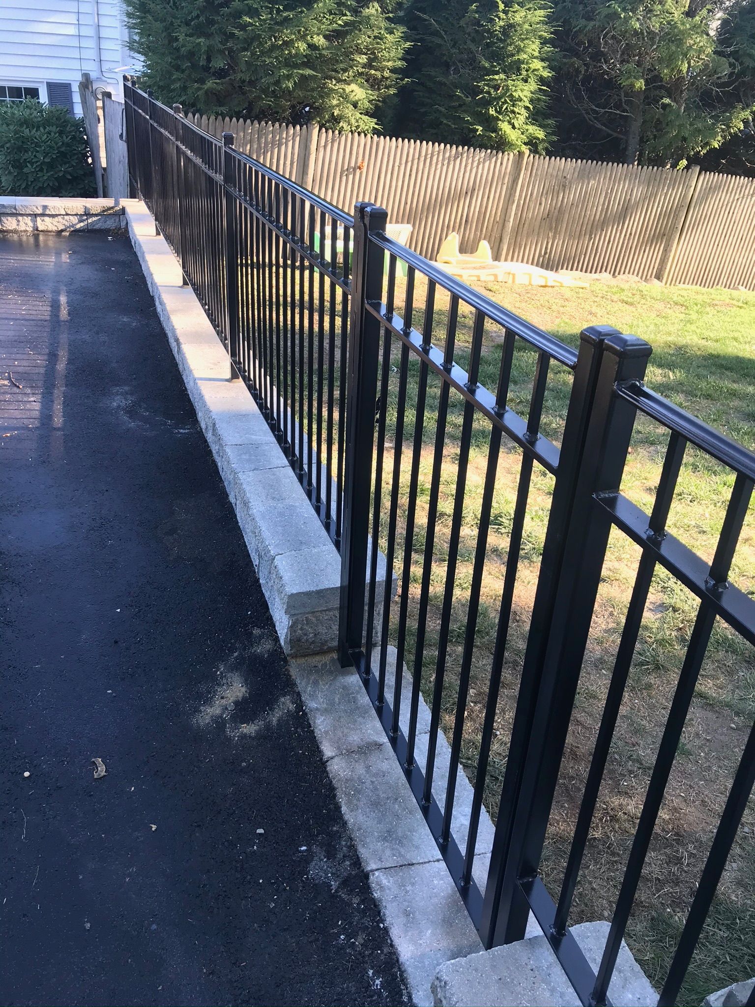 Black metal fence along a concrete edge beside a paved driveway and grassy yard with a wooden privacy fence.
