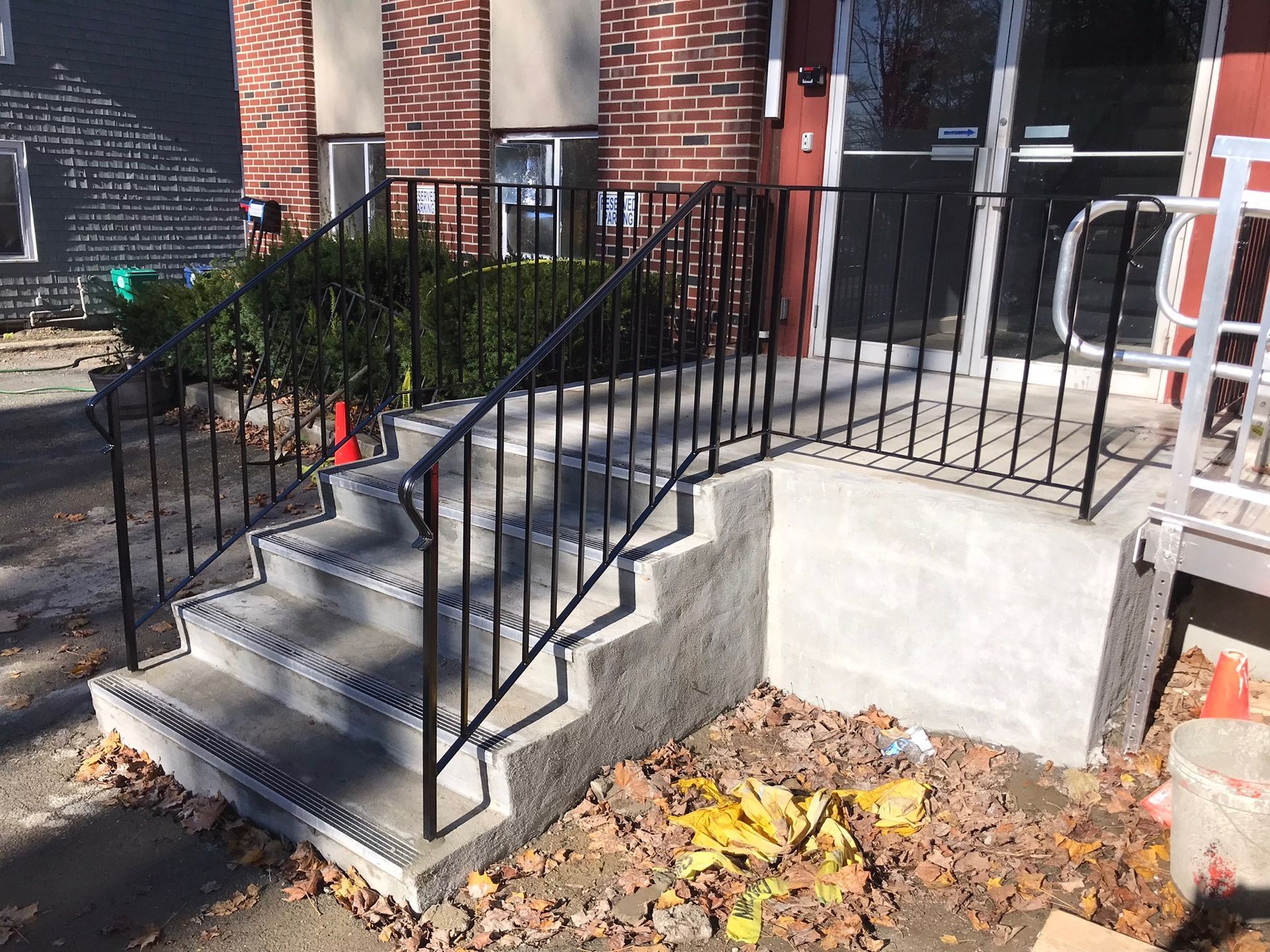 Front steps with black railings leading to a brick building entrance, bordered by a concrete ramp and autumn leaves.