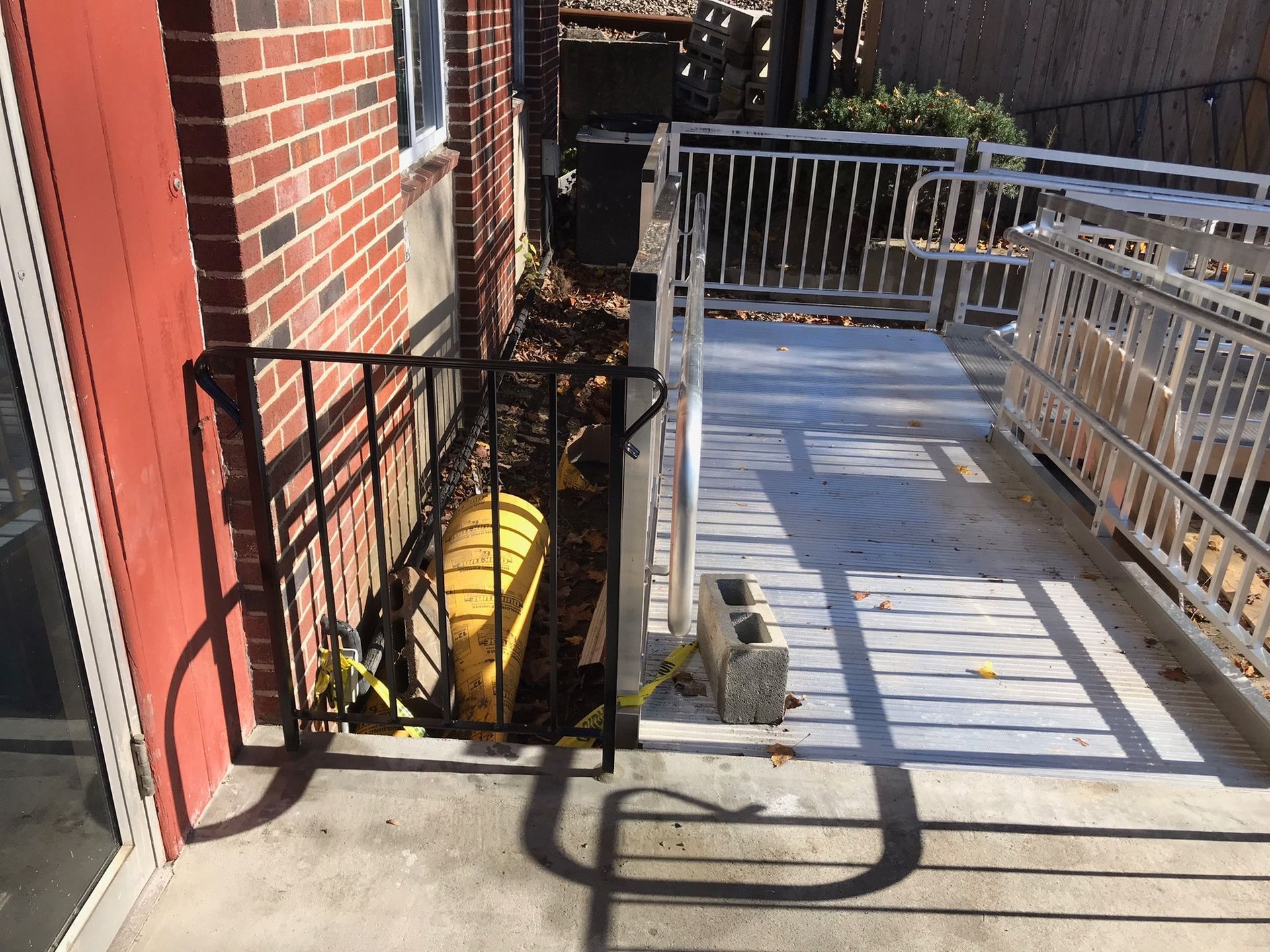 Narrow backyard walkway beside a brick house, with black railing, ramp shadows, and a yellow hose reel.