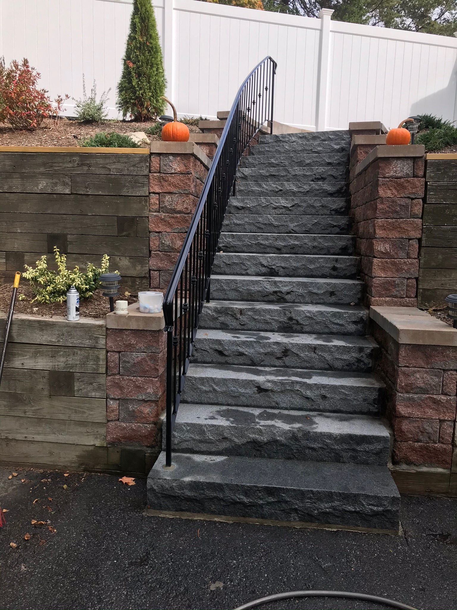 Steep outdoor stone staircase with black handrails leading up to a white fence, flanked by brick retaining walls and pumpkins.