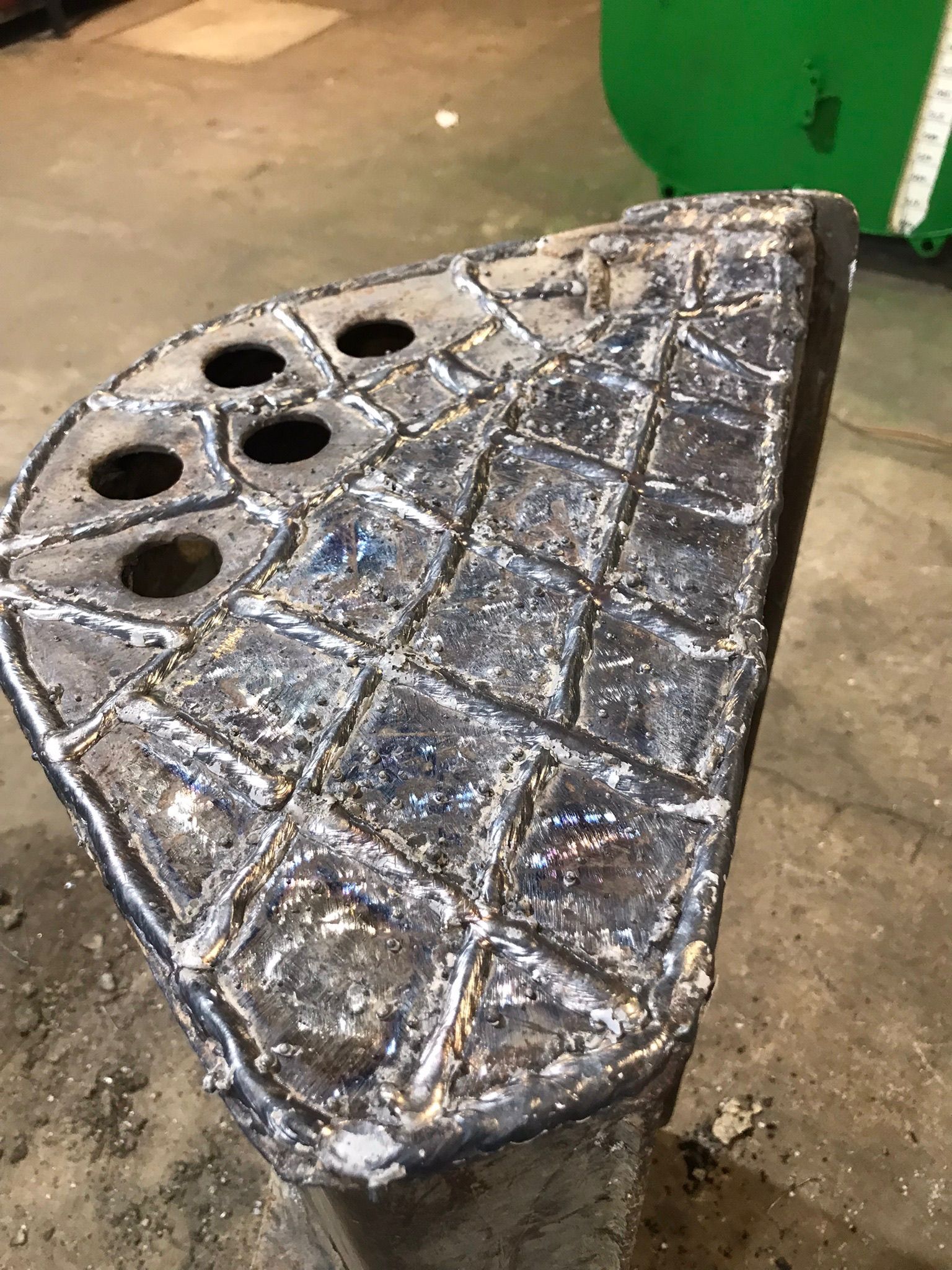 Worn metal triangular plate with several circular holes, lying on a concrete floor in a workshop.